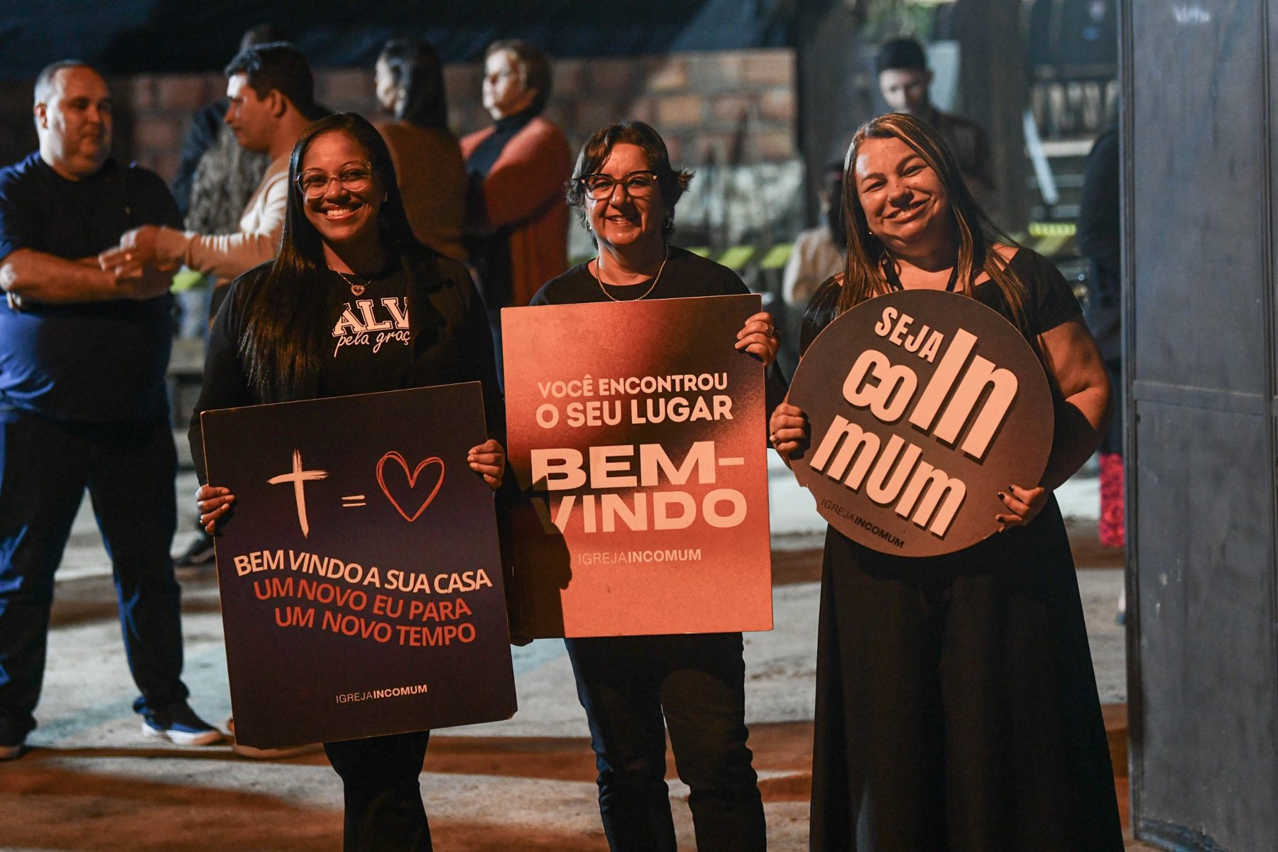 Três mulheres da recepção sorrindo com placas de boas-vindas da Igreja Incomum